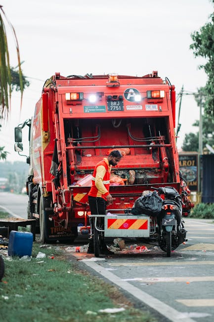 A worker dressed in a yellow and red uniform is operating a red rubbish collection vehicle parked on the side of a road, with the vehicle's rear open for waste loading. The worker is focused on the area beneath the open compartment, which contains debris and waste materials. In front of the vehicle, a small black motorcycle with a black trash bag or container attached is positioned, possibly for additional waste collection. The surrounding environment includes a stretch of road, some greenery and trees in the background, and utility poles with power lines overhead. The overall scene depicts on-site rubbish clearance, highlighting a professional waste management process outside an urban setting, consistent with private waste handling services. The vehicle appears to be in good condition, with clear markings and lights, supporting the context of independent rubbish removal, often used in situations where local authority collection is not available or preferred. The lighting is natural, suggesting daytime with overcast skies, and the scene emphasizes the practical aspect of rubbish disposal in a professional waste clearance operation.