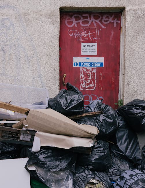 A worker dressed in a yellow and red uniform is operating a red rubbish collection vehicle parked on the side of a road, with the vehicle's rear open for waste loading. The worker is focused on the area beneath the open compartment, which contains debris and waste materials. In front of the vehicle, a small black motorcycle with a black trash bag or container attached is positioned, possibly for additional waste collection. The surrounding environment includes a stretch of road, some greenery and trees in the background, and utility poles with power lines overhead. The overall scene depicts on-site rubbish clearance, highlighting a professional waste management process outside an urban setting, consistent with private waste handling services. The vehicle appears to be in good condition, with clear markings and lights, supporting the context of independent rubbish removal, often used in situations where local authority collection is not available or preferred. The lighting is natural, suggesting daytime with overcast skies, and the scene emphasizes the practical aspect of rubbish disposal in a professional waste clearance operation.