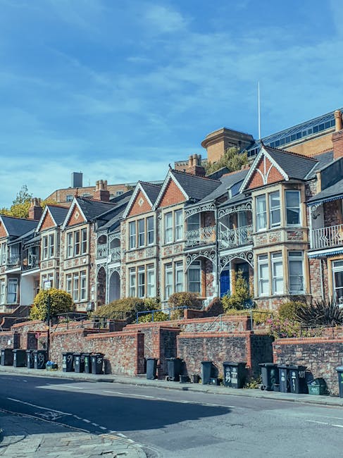A row of Victorian-style terraced houses with ornate white decorative trim on the facades, featuring bay windows and small balconies, set above a brick wall with planting beds and shrubs in front. The buildings are constructed from red brick with contrasting white accents and dark roof tiles. The scene is illuminated by daylight with a partly cloudy sky overhead, and a street with black wheelie bins lined up along the pavement in the foreground. Visible behind the houses are modern buildings with glass and concrete elements, indicating an urban setting. This image relates to private waste handling options such as independent rubbish collection or on-site clearance, reflecting the local residential environment. The overall setting appears calm and tidy, with no visible signs of debris or rubbish in the public space, highlighting the importance of organized waste management services provided by companies like Waste Clearance Balham.