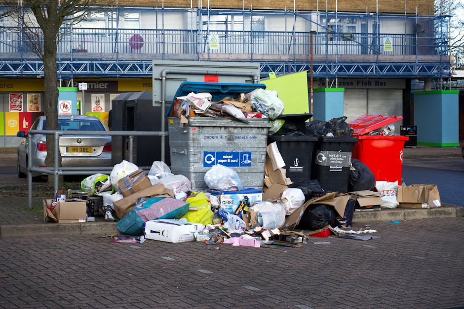 A collection of overflowing rubbish and waste materials accumulated around multiple refuse bins on a paved area in an urban setting. The waste includes cardboard boxes, plastic bags, crumpled papers, and miscellaneous household refuse, some spilling onto the ground. The central bin is a large, grey, wheeled container designated for mixed paper and cardboard, partially open with flattened boxes and loose trash visible inside. Surrounding it are black and red bins. In the background, a blue construction scaffold covers a building facade with blue metal fencing on top, and storefronts with signs and advertisements are visible, indicating commercial units nearby. Vehicles are parked adjacent to the waste collection area, including a silver car with the license plate partially visible. The scene is lit with natural daylight, highlighting the disorganized state of the waste. This image relates to private rubbish removal services that provide alternative waste handling options away from council disposal, aligning with the purpose of waste clearance professionals like Waste Clearance Balham.