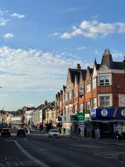 A row of Victorian-style terraced houses with ornate white decorative trim on the facades, featuring bay windows and small balconies, set above a brick wall with planting beds and shrubs in front. The buildings are constructed from red brick with contrasting white accents and dark roof tiles. The scene is illuminated by daylight with a partly cloudy sky overhead, and a street with black wheelie bins lined up along the pavement in the foreground. Visible behind the houses are modern buildings with glass and concrete elements, indicating an urban setting. This image relates to private waste handling options such as independent rubbish collection or on-site clearance, reflecting the local residential environment. The overall setting appears calm and tidy, with no visible signs of debris or rubbish in the public space, highlighting the importance of organized waste management services provided by companies like Waste Clearance Balham.