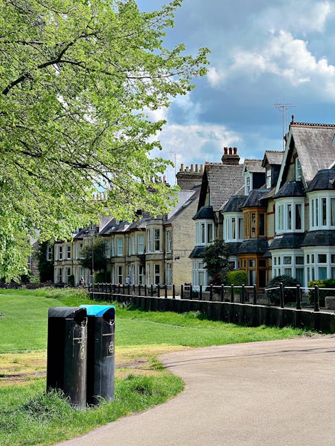 A row of traditional terraced houses with bay windows, brickwork in light beige and darker shades, and chimneys visible on the rooftops along a residential street. In the foreground, a wide, paved pathway runs parallel to the houses, with lush green grass and a black metal fence separating the properties from the pavement. To the left, a large deciduous tree with bright green foliage extends its branches across the scene, casting partial shade over the pathway and grass. Near the bottom left, two black cylindrical waste bins with blue lids are positioned on the grass, with one bin slightly leaning. The sky above is partly cloudy with patches of blue visible through the white clouds, suggesting a bright, clear day. This scene reflects a quiet residential area where private waste collection or informal rubbish disposal might be located near the homes, consistent with services offered by waste clearance specialists such as Waste Clearance Balham.