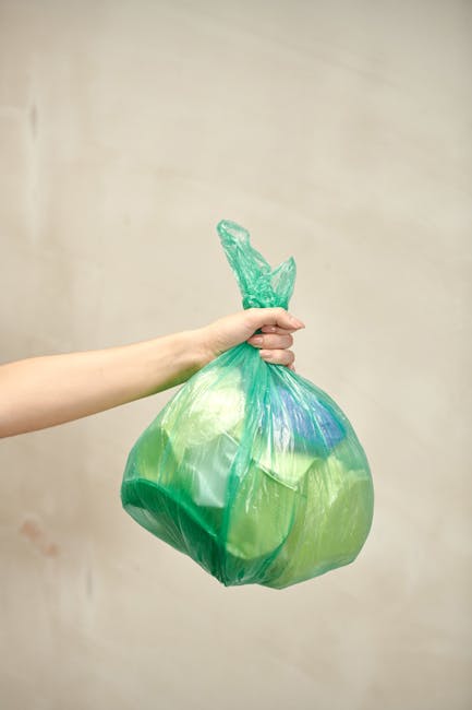 A person’s hand and forearm extend from the left side of the image, holding a green plastic rubbish bag by its twisted top knot. The bag is semi-transparent, revealing an assortment of waste materials inside, and has a smooth, slightly crinkled texture. It appears filled with light debris, giving it a rounded, bulging shape. The background features a plain, neutral-toned wall with a subtle gradient, giving an unobtrusive backdrop that emphasizes the bag's vivid green colour. The scene is lit with soft, even lighting, highlighting the bag's glossy surface and the hand’s natural skin tone. This visual collectively depicts a typical scene of waste disposal, aligning with the context of rubbish removal services such as those offered by Waste Clearance Balham, emphasizing the importance of private collection or on-site clearance alternatives for household waste. The image underscores the act of removing waste from domestic or commercial settings, relevant for an article discussing rubbish collection options near Balham Station or similar urban locations.