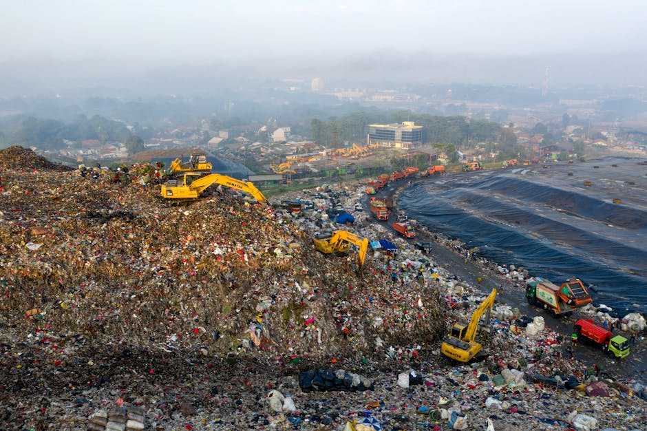 An expansive landfill site filled with a large mound of mixed waste materials, primarily consisting of plastic bottles, bags, and other household refuse. Several yellow excavators are actively engaged in moving and sorting the rubbish on the uneven, debris-covered surface. To the right, a section of the waste area is covered with black plastic sheeting, possibly for containment or covering older waste. In the distance, there is a row of trucks and vehicles parked along a dirt or gravel road that borders the dump, and some green waste collection vehicles are visible among them. Beyond the landfill, the background shows a hazy cityscape with numerous low-rise buildings, trees, and a soft, overcast sky, contributing to a somber, utilitarian atmosphere typical of waste management and disposal sites. This scene exemplifies the type of infrastructure associated with independent rubbish and waste clearance services, highlighting the scale of material handling involved in refuse disposal.