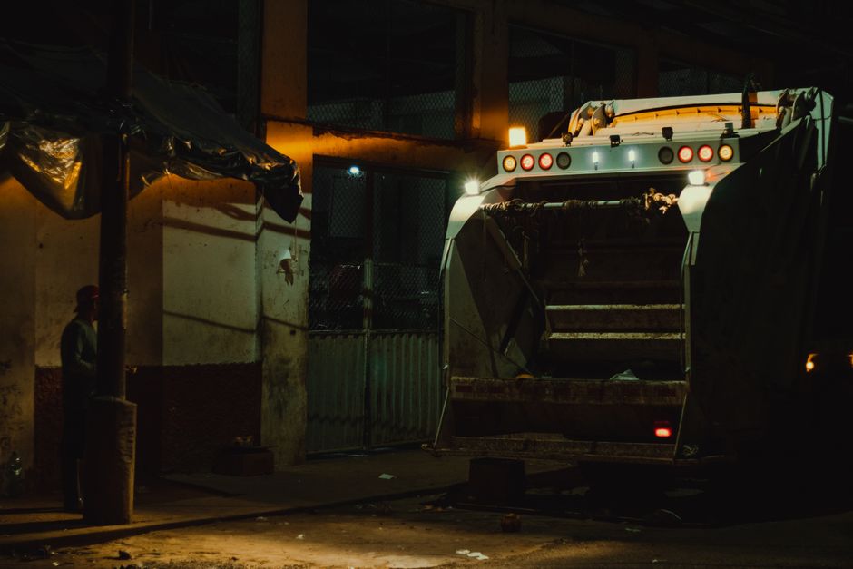 A large waste collection truck with an open rear hopper is parked outdoors at night under dim lighting, revealing a predominantly white and gray metal body with multiple circular red and white warning lights on the top. The truck's cargo area appears empty or contains minimal load, with visible dirt and debris on the metal surfaces. The scene is illuminated by nearby artificial lights, casting shadows on the surrounding environment, which includes a concrete wall on the left side with a partially covered section using dark tarpaulin, and a metal gate or fence behind the truck. To the left, a person wearing a high-visibility vest and a hard hat stands next to a wooden pole, observing the area. The setting suggests an industrial or commercial site involved in waste or rubbish collection, possibly during a private or on-site clearance operation managed by Waste Clearance Balham, with minimal natural light and a focus on the operational equipment for rubbish removal tasks.